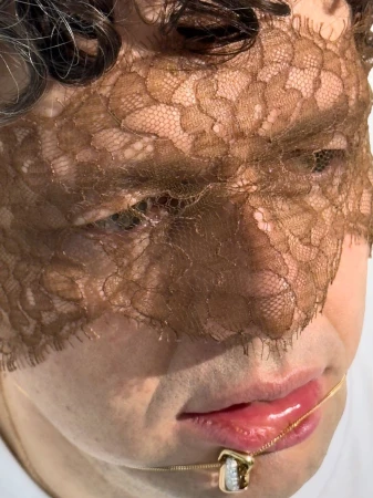 A close-up of a person's face partially covered with lace, emphasizing a gold necklace and soft lighting.