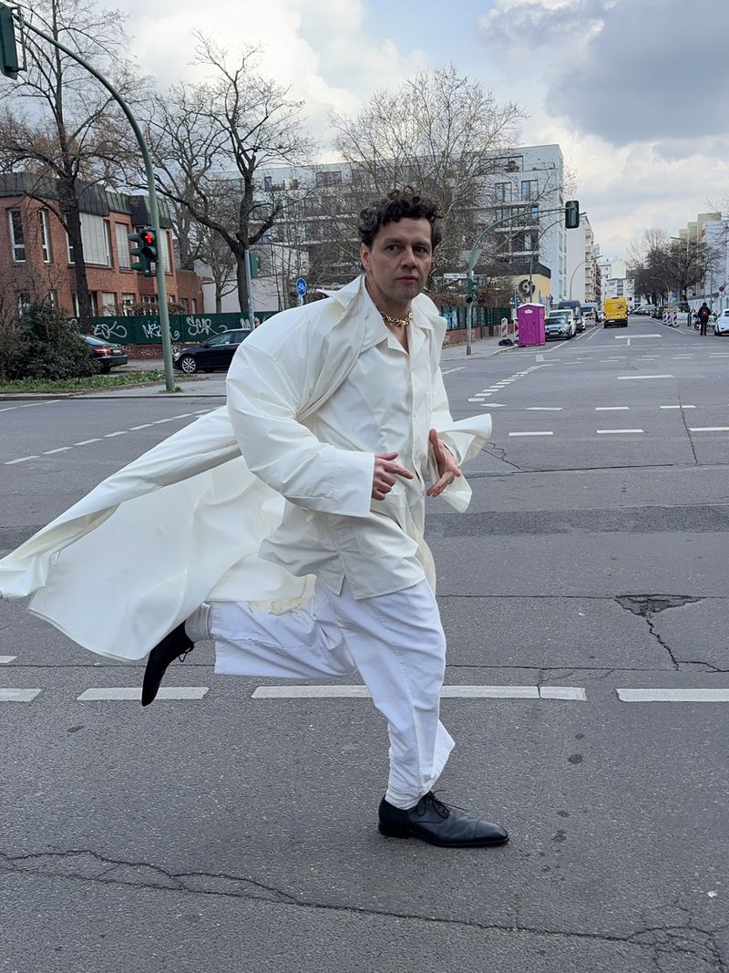 Man in white coat and pants runs across a city street with buildings and traffic lights visible under a cloudy sky during daytime.