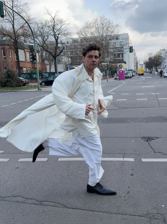Man in white coat and pants runs across a city street with buildings and traffic lights visible under a cloudy sky during daytime.
