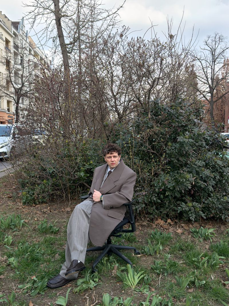 Man in oversized suit sits on office chair outdoors surrounded by green grass and foliage. Building visible in background.