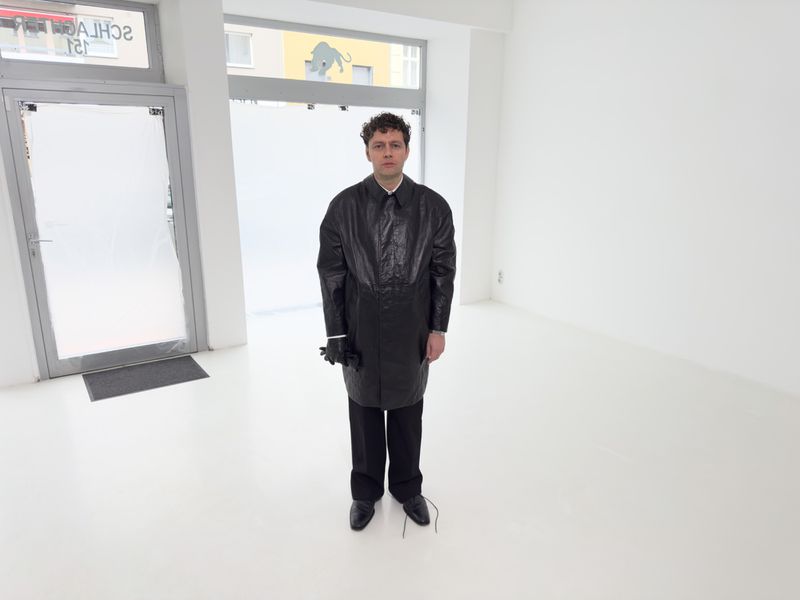 Man in a black coat stands in an all-white room with matte floors, wearing gloves and dress shoes, near a frosted glass door.