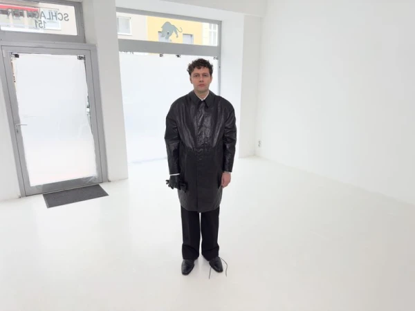 Man in a black coat stands in an all-white room with matte floors, wearing gloves and dress shoes, near a frosted glass door.