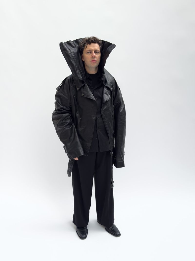Full shot of a person with curly hair wearing an oversized black leather jacket and wide-legged trousers against a plain white backdrop.