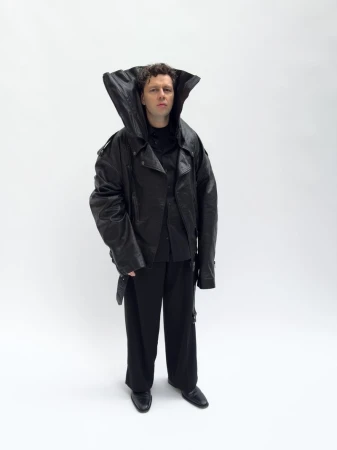 Full shot of a person with curly hair wearing an oversized black leather jacket and wide-legged trousers against a plain white backdrop.