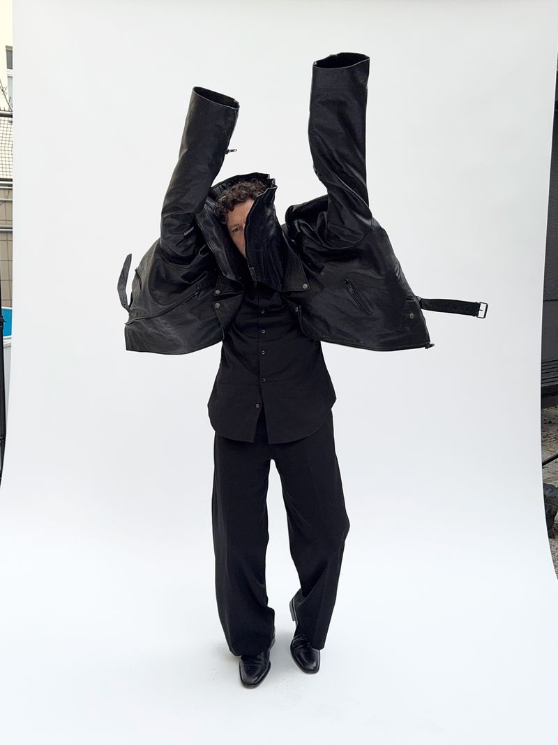 Fashionable figure strikes a pose in an all-black ensemble, dramatically holding a black jacket aloft against a stark white backdrop.