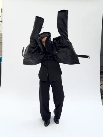 Fashionable figure strikes a pose in an all-black ensemble, dramatically holding a black jacket aloft against a stark white backdrop.