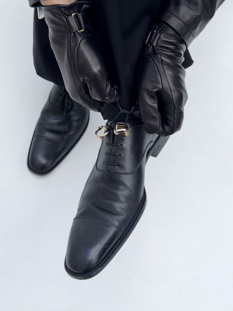 A close-up shot of a person in black gloves tying the laces of polished black leather dress shoes, set against a stark white background.