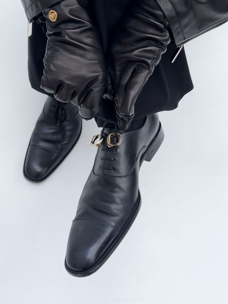 Close-up shot of a man adjusting the decorative gold buckles on his black leather dress shoes, complemented by black leather gloves and jacket.