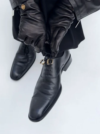 Close-up shot of a man adjusting the decorative gold buckles on his black leather dress shoes, complemented by black leather gloves and jacket.