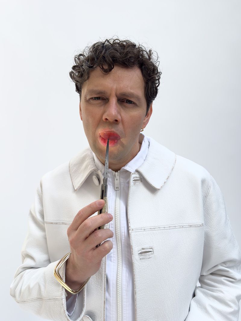 Adult male holding a knife in his mouth, posing in a white jacket against a white backdrop, with an intense expression. Studio shot.