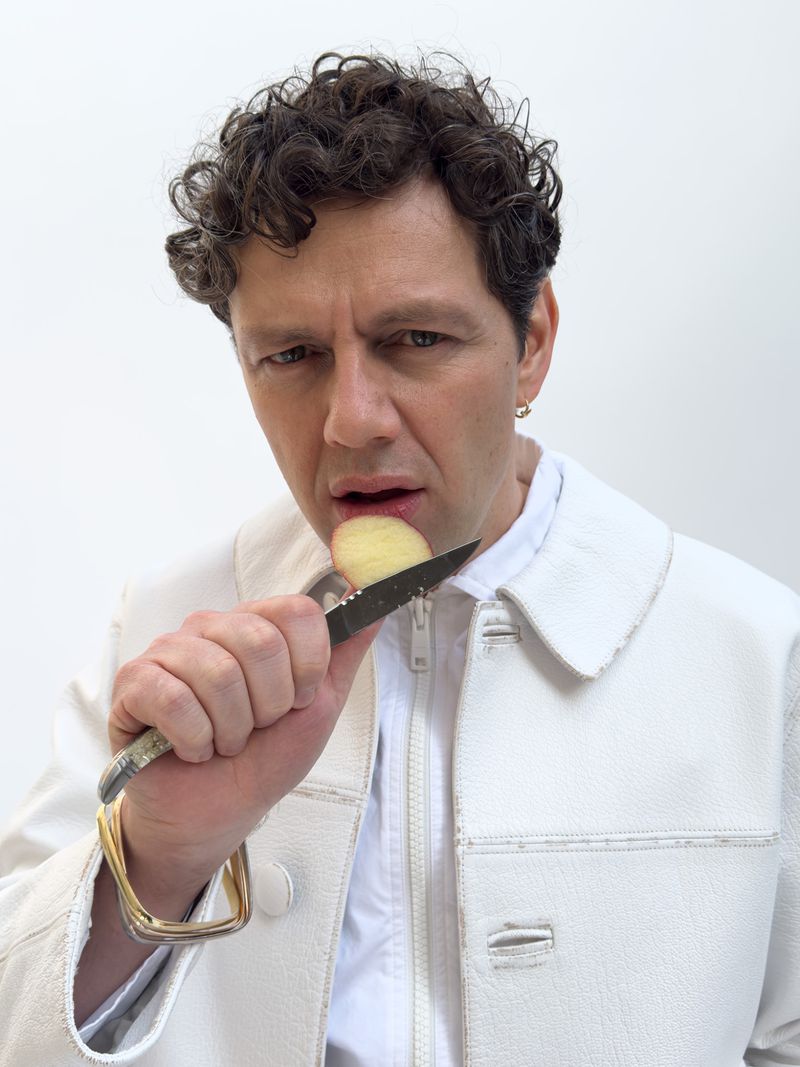 Portrait of a man holding a knife with a potato on the blade, wearing a white jacket, set against a plain white background.