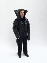 Man wearing an oversized black leather jacket with a high structured collar against a plain white background