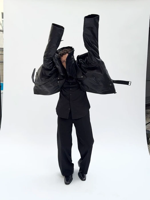 Christian Friedel posing with an oversized black leather jacket lifted over his head against a white seamless background.