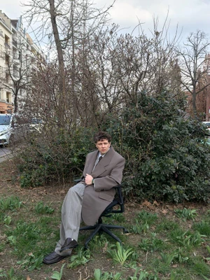 Actor Christian Friedel seated in an office chair outdoors in a taupe overcoat and pinstripe trousers against bushes.