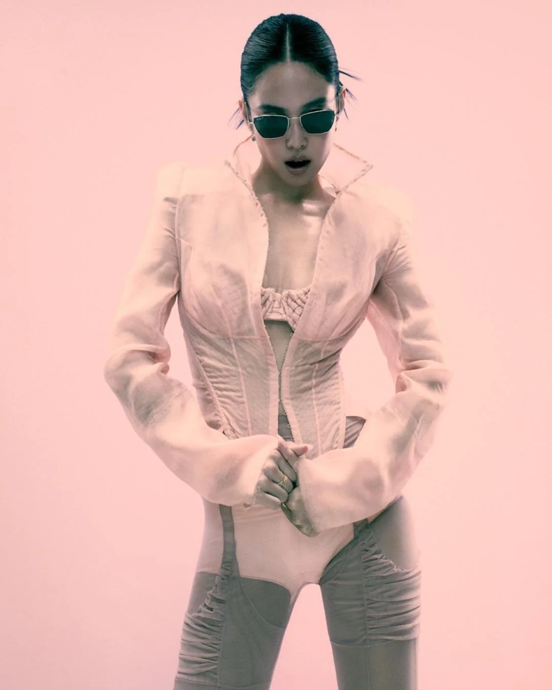 Jennie posing in dark sunglasses and a structured pink corset jacket against a soft pale pink backdrop.