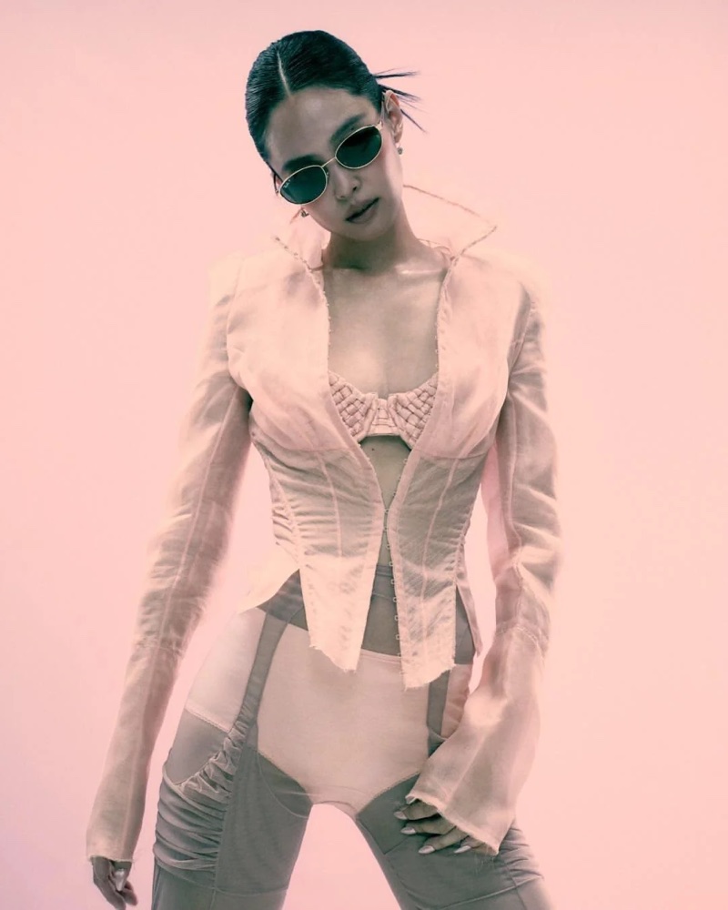 Jennie wearing dark oval sunglasses and an open sheer pale pink jacket against a pink studio backdrop.