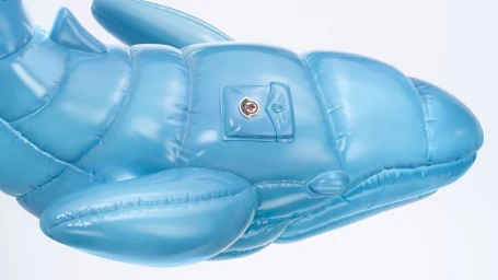 Close-up of a light blue inflatable puffer whale sculpture with quilted texture and a Moncler logo pocket against a plain white background