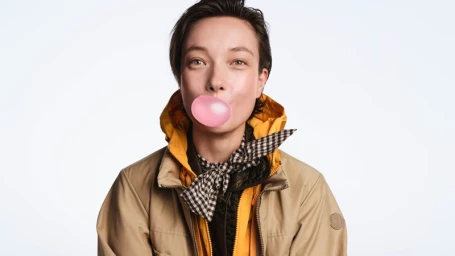 Yumi Lambert wearing a yellow jacket and checkered scarf while blowing a pink bubblegum bubble against white background
