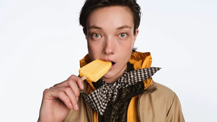 Model Yumi Lambert wearing a yellow jacket and checkered scarf while eating an orange ice pop on a white background for Moncler.