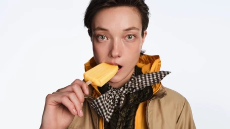 Model Yumi Lambert wearing a yellow jacket and checkered scarf while eating an orange ice pop on a white background for Moncler.