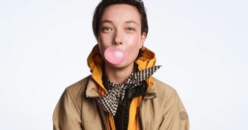 Model Yumi Lambert blowing a pink bubble gum bubble wearing a khaki jacket and checkered scarf against a white background for Moncler Puffy Summer 2026