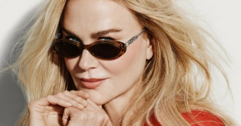 Close-up portrait of Nicole Kidman wearing brown tortoiseshell sunglasses and a red top with wind-swept blonde hair against a white background