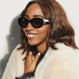 Ayo Edebiri smiling while wearing black sunglasses and a textured white fur-style coat against a pale wall.