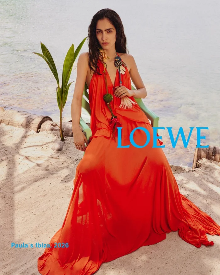 Model Sara Caballero in a vibrant orange dress seated outdoors on a sandy beach for Loewe Paula's Ibiza 2026.