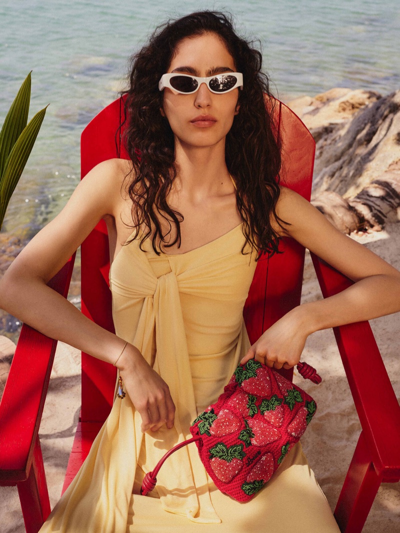 Model Sara Caballero sitting in a red chair on a sandy beach wearing a yellow dress, white sunglasses, and holding a strawberry-patterned bag.
