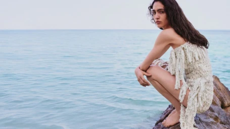 Model Sara Caballero in a patterned white dress crouching on coastal rocks by the ocean for Loewe Paula's Ibiza 2026