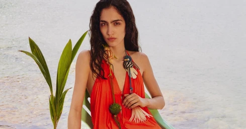 Sara Caballero in a red halter dress with woven bird accessories seated outdoors by the water for Loewe Paula's Ibiza 2026