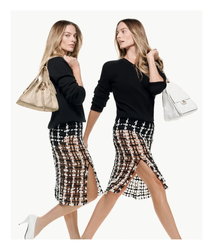 Margot Robbie in a black sweater and tweed skirt holding a Chanel handbag against a white background.