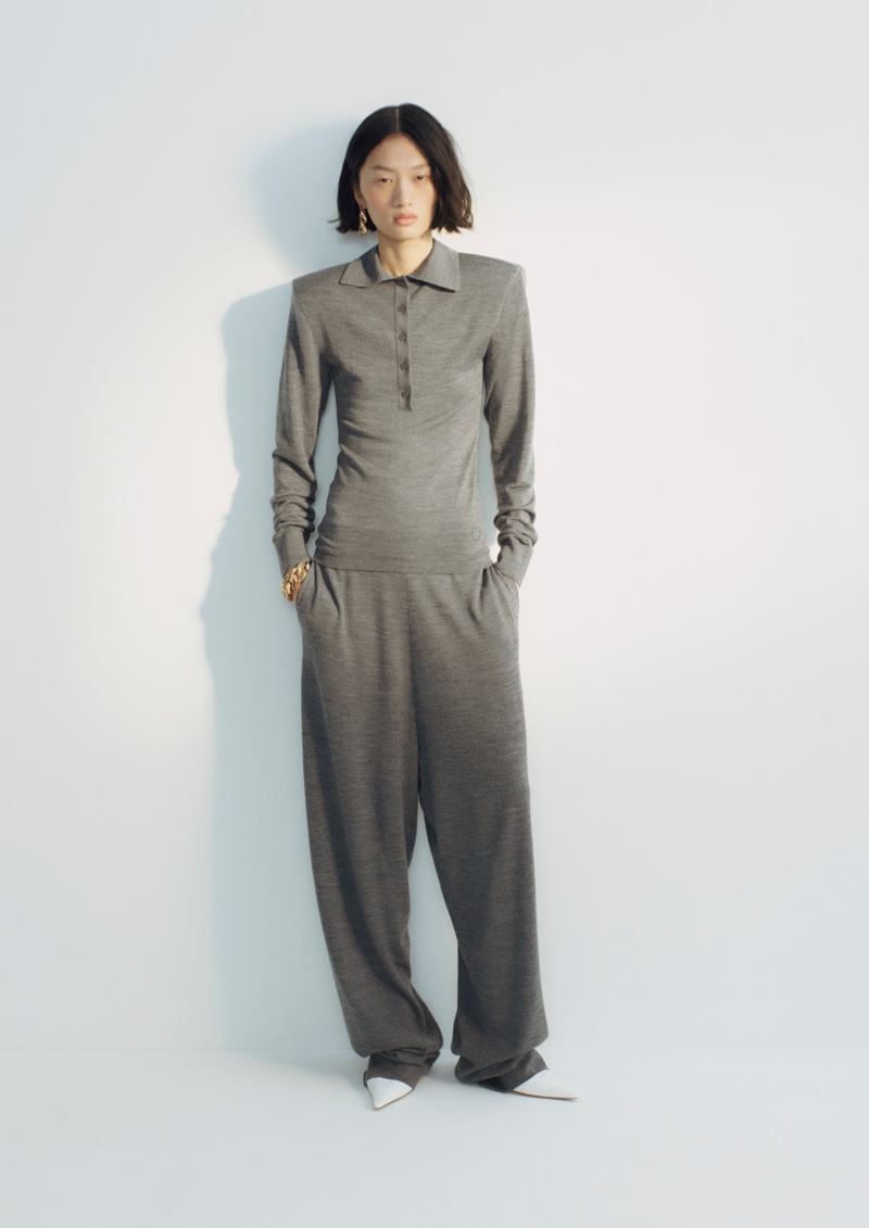 Model Angelina Kendall in a grey long-sleeve polo sweater and matching loose trousers against a white studio background.