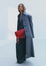 Model Angelina Kendall in a long grey coat holding a red bag against a white studio background