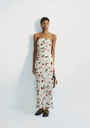 Model standing in a strapless cream maxi dress with cherry print and black chain loafers against a white studio background.