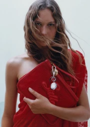 Model holding a red zippered pouch bag with metal charms wearing a red dress against a plain light background