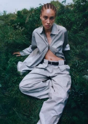 Model Adwoa Aboah seated in a lush grassy meadow wearing a light grey matching button-down shirt and trousers set by H&M and Stella McCartney.