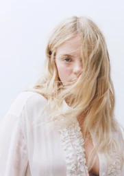 Model with long blonde hair partially covering her face wearing a sheer white ruffled shirt against a white background.