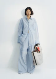 Model in oversized blue striped shirt and matching trousers holding white handbags against a white backdrop