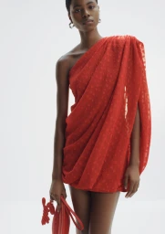 Black model wearing an asymmetrical red dress holding a red clutch against a white background