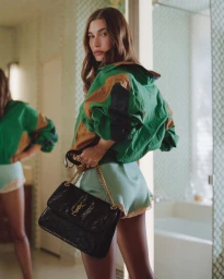 Hailey Bieber wearing green jacket and holding a black handbag in a domestic interior by Nadia Lee Cohen for Yves Saint Laurent.