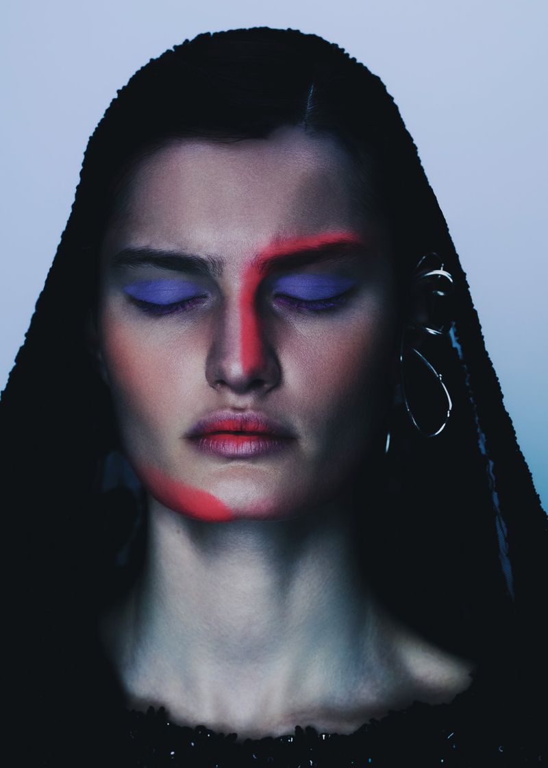 Close-up beauty portrait of a woman with eyes closed featuring red and purple face makeup, silver ear jewelry, and a black head covering.