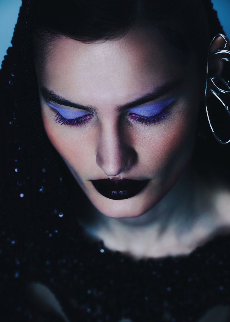 Close-up portrait of a woman with bold purple eye makeup, dramatic black lipstick, and a sequined top against a cool-toned background.