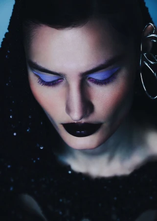 Close-up portrait of a woman with bold purple eye makeup, dramatic black lipstick, and a sequined top against a cool-toned background.