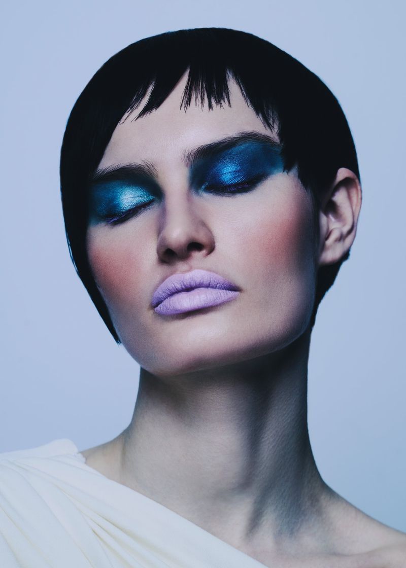 Close-up portrait of a female model with closed eyes, wearing blue eyeshadow, purple lipstick, and a white top against a light blue background.