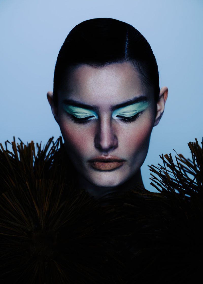 Close-up studio shot of a woman with short dark hair, light green eyeshadow, and a dark, feathered collar against a light blue background.