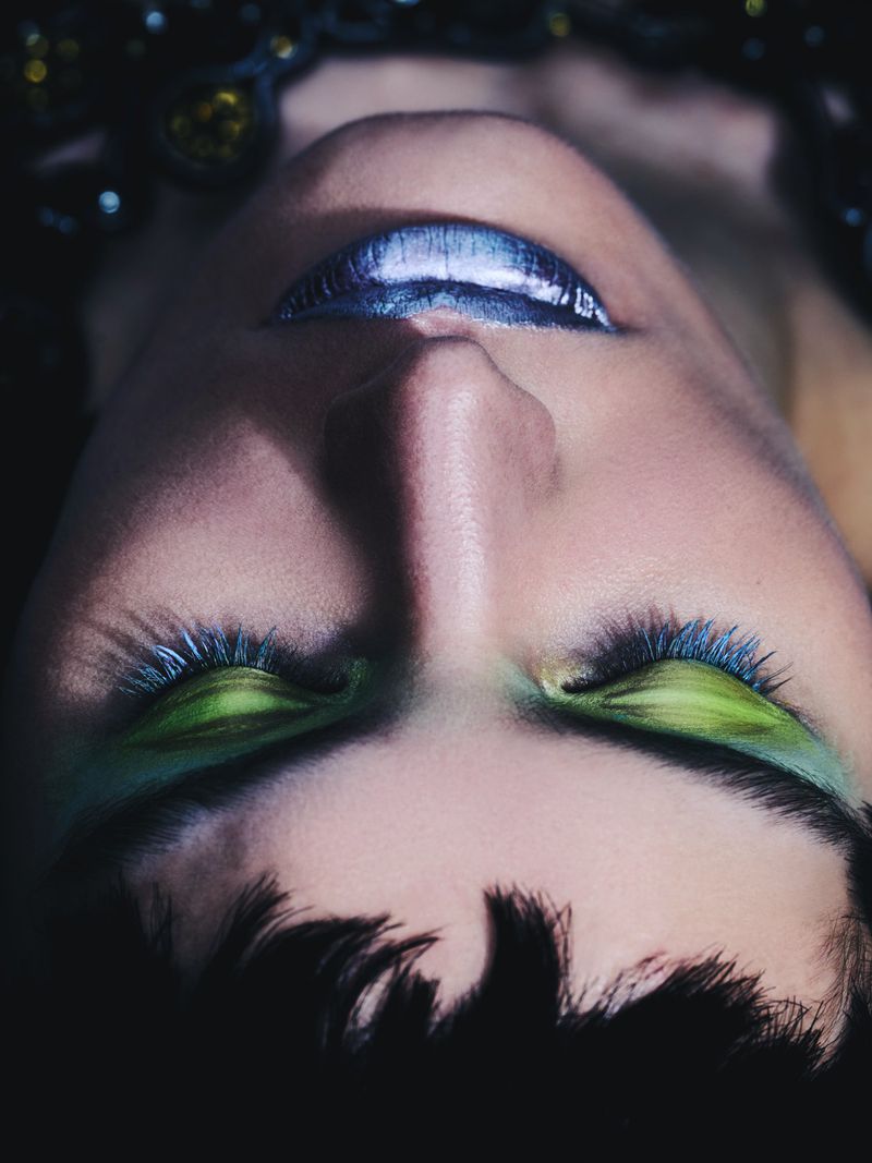 Close-up studio shot of an adult woman with blue lipstick and green eyeshadow, captured with dramatic, dark lighting and eye-level angle.