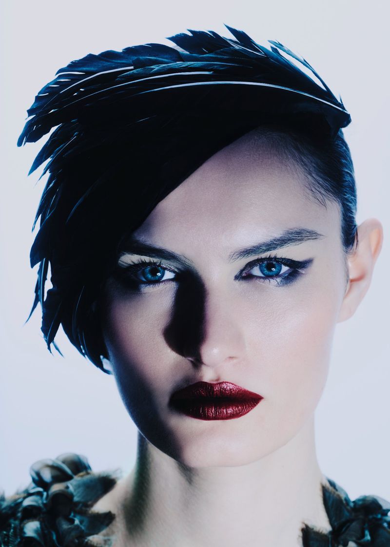 Portrait of a woman with dramatic makeup and a black feather headdress against a bright backdrop in a studio setting.