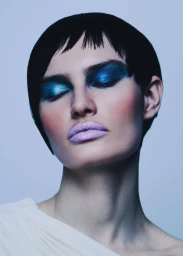Close-up portrait of a woman with short dark hair, striking metallic blue eyeshadow, and pale lavender lipstick against a plain background