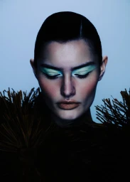 Close-up portrait of a model with green eyeshadow and textured brown collar against a pale blue background
